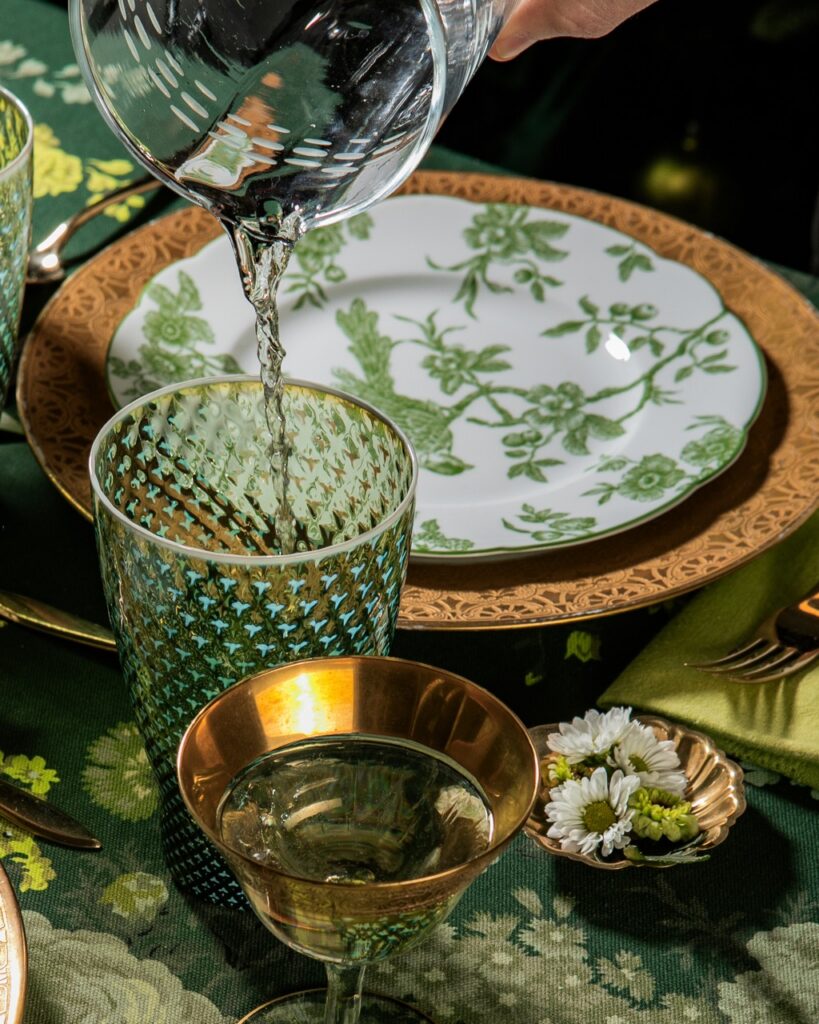 A person pours water into a green glass with a white and green and brown plate in the background.