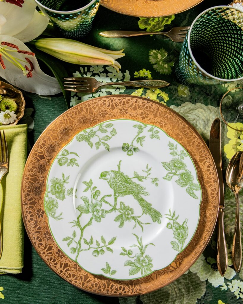 A white plate with a green bird pattern on it sits on a table setting with gold embellishments.
