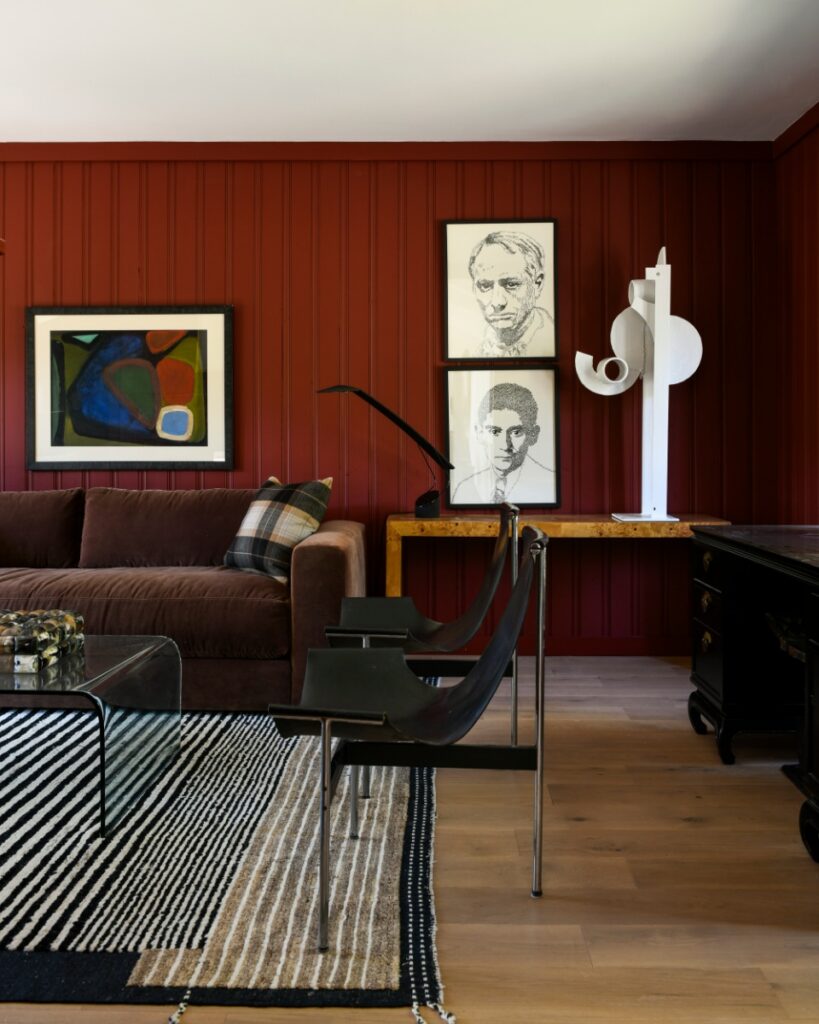 A Fox Chapel den featuring deep red walls, a vintage wooden console table, a brown velvet sofa, and a vibrant geometric patterned area rug.