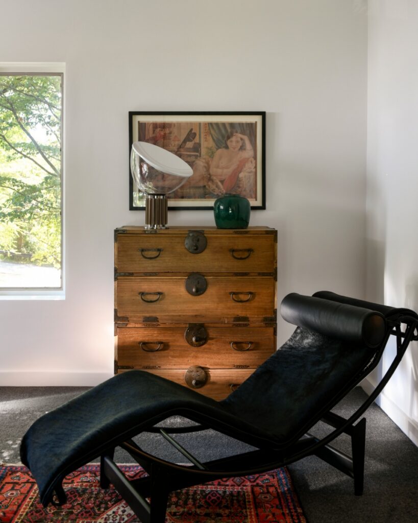 A primary bedroom corner in Fox Chapel featuring a black leather chaise longue, a vintage wood tansu chest, a green ceramic vase, and a Flos Taccia table lamp.