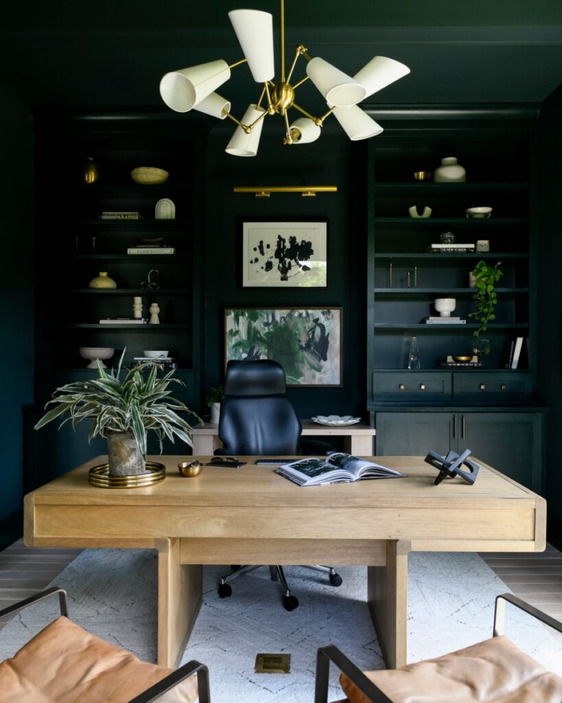A moody, dark green home office in a Fox Chapel residence featuring floor-to-ceiling built-in shelving, a light wood desk, and a sculptural cream-colored chandelier designed by Elysa Roberts Interiors.
