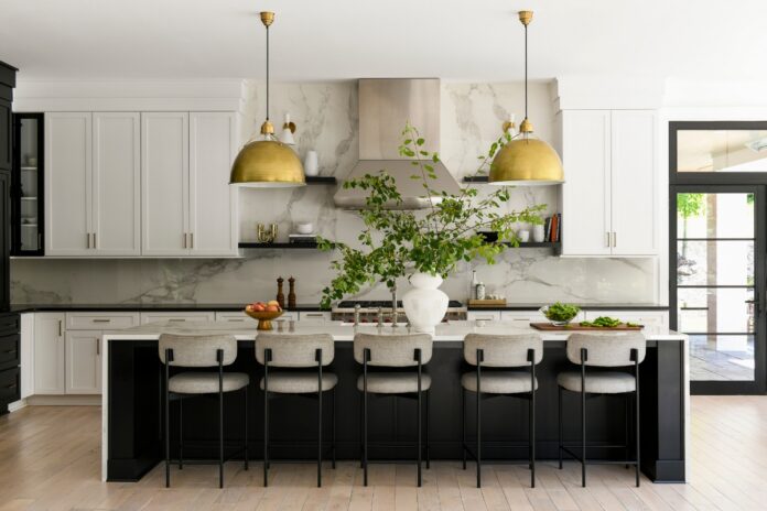 A bright California-cool kitchen in a Fox Chapel home featuring a large black island with a marble countertop, brass pendant lights, and white cabinetry designed by Elysa Roberts Interiors.