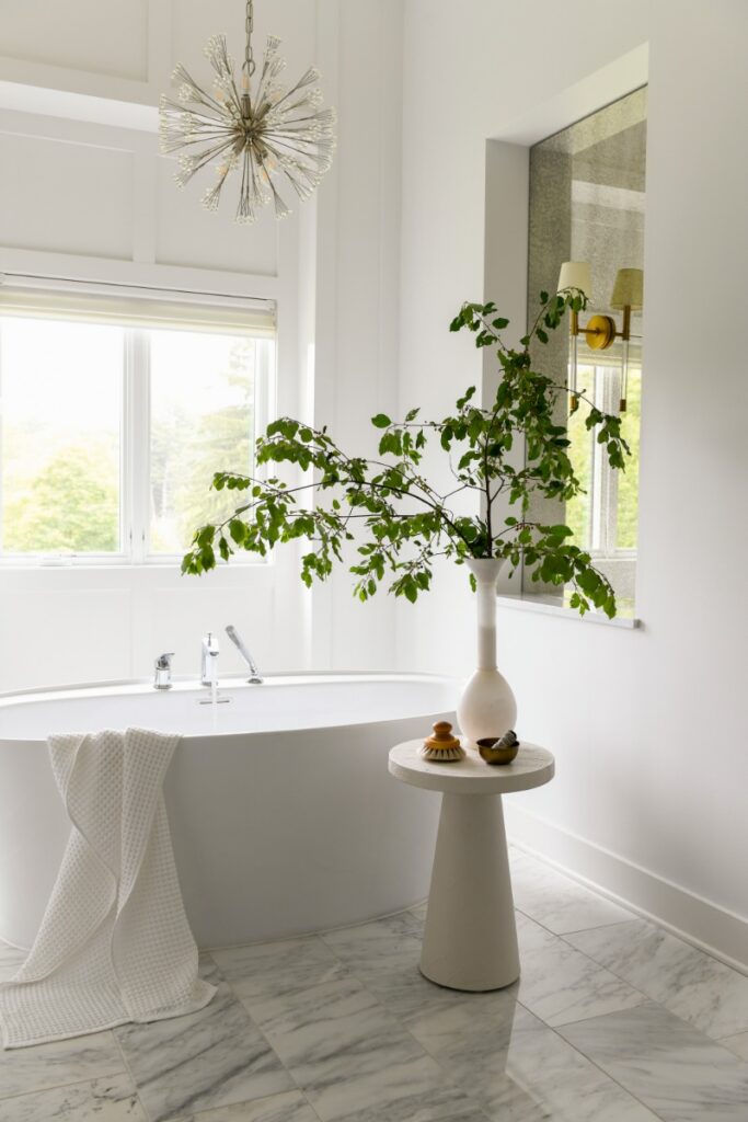 A light-filled primary bathroom in a Fox Chapel home featuring a freestanding white soaking tub, marble tile flooring, and a modern sputnik-style chandelier.