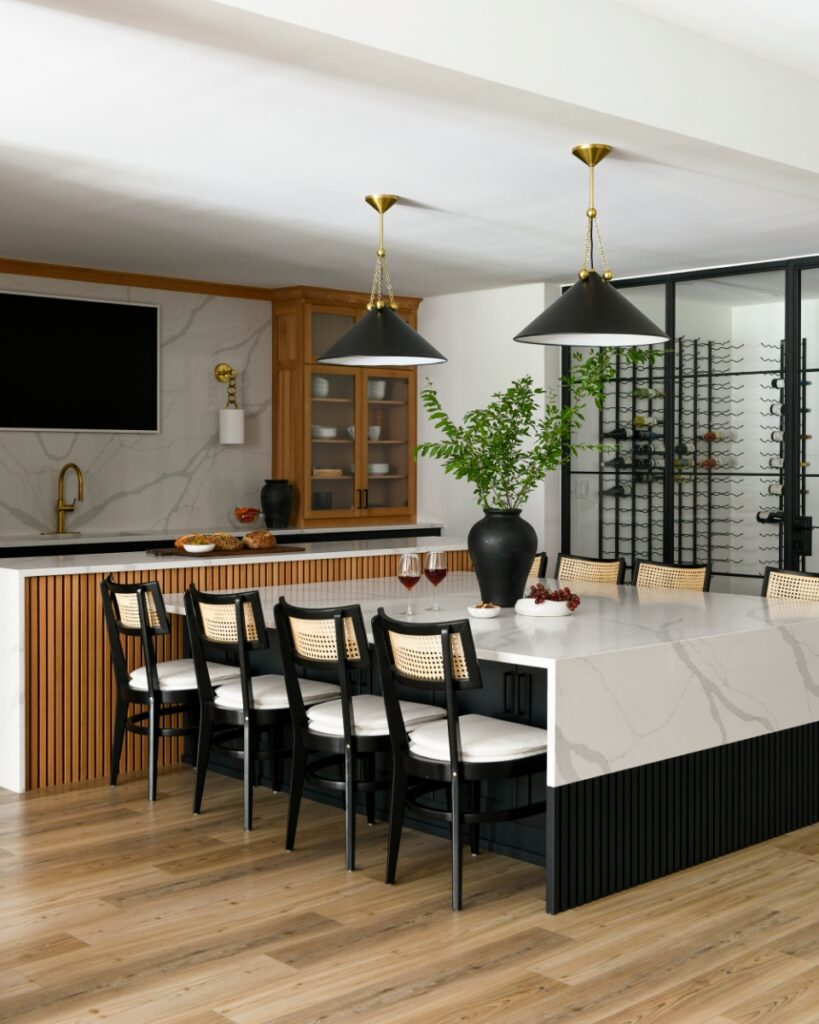 A contemporary basement bar and kitchen featuring a large marble island, black fluted cabinetry, and a floor-to-ceiling glass wine cellar designed by Elysa Roberts Interiors.