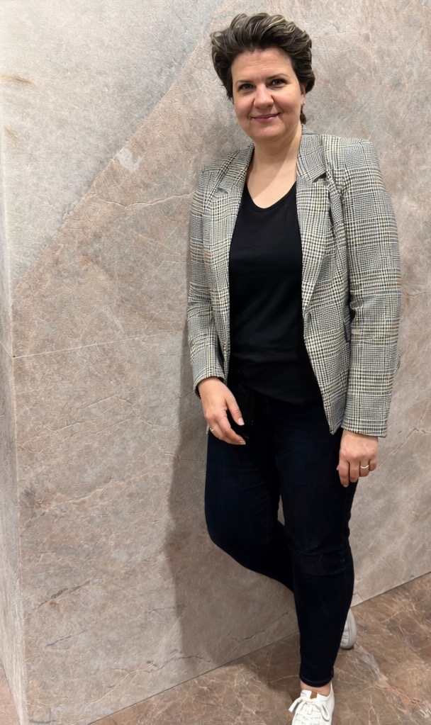 A woman stands in a grey blazer and black clothing with short hair.