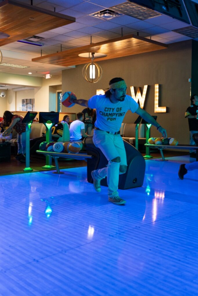 A person in a "City of Champyinz" t-shirt bowling on a neon-lit blue lane at AMF Mt. Lebanon Lanes.