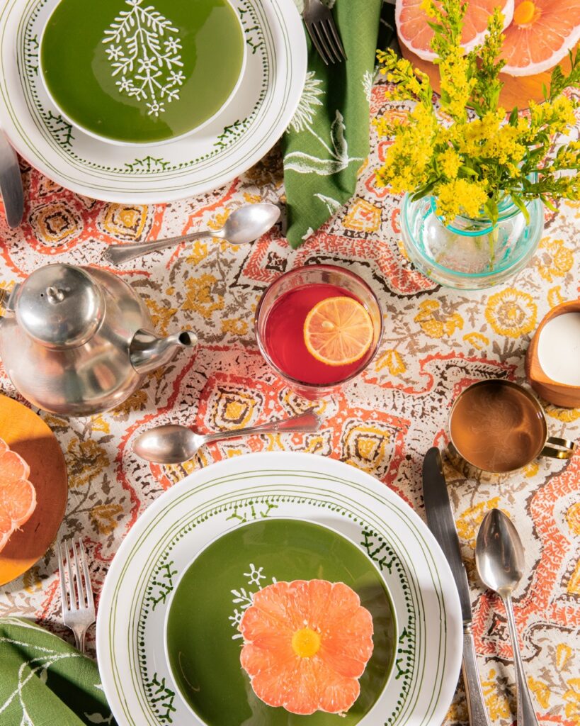 Two plate settings with stacks of white and green plates on an orange table cloth and glasses of drinks.