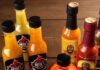 Pittsburgh Hot Sauces Made Locally for the Grilling Season A variety of Pittsburgh-made local hot sauces from Artie’s, Uncle Jammy’s, and Bazaaa Sauce arranged on a wooden table with fresh habanero and Fresno peppers.