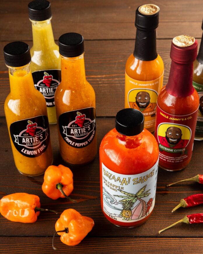 A variety of Pittsburgh-made local hot sauces from Artie’s, Uncle Jammy’s, and Bazaaa Sauce arranged on a wooden table with fresh habanero and Fresno peppers.