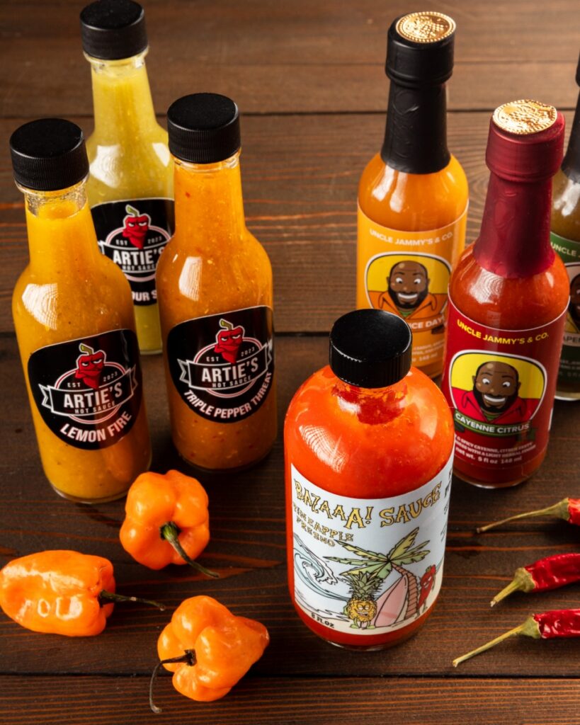 A variety of Pittsburgh-made local hot sauces from Artie’s, Uncle Jammy’s, and Bazaaa Sauce arranged on a wooden table with fresh habanero and Fresno peppers.