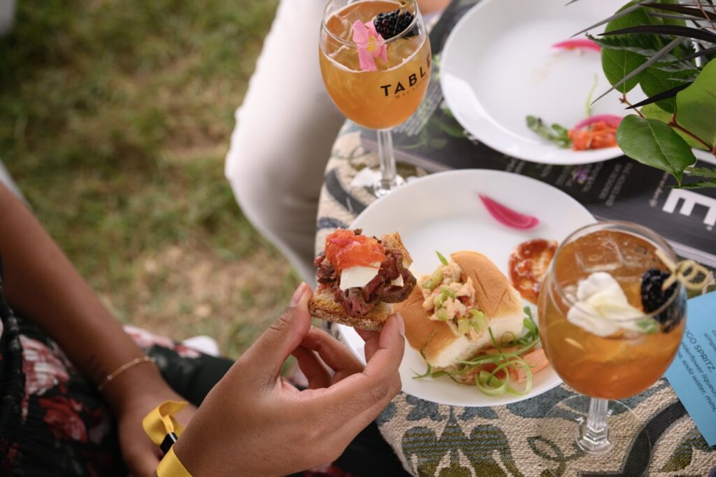 A person picks up a small appetizer from a plate of food between two cocktails.