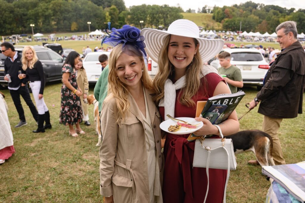 Two women smile in a fascinator and large hat.