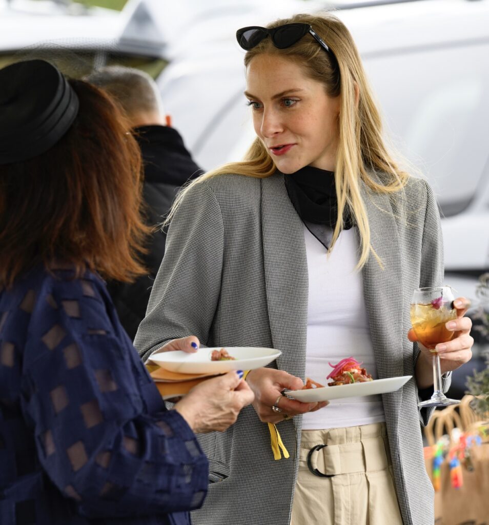 A woman talks to another with a plate of food and cocktail glass in hand.
