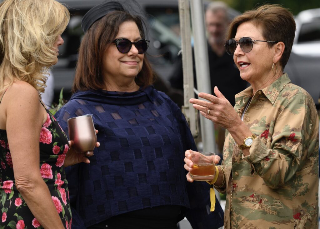 Three women talk with cocktails in their hands and sunglasses on.