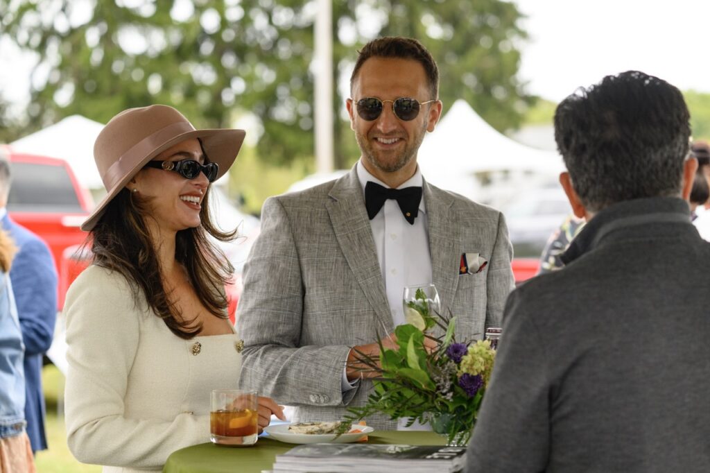 A man in a suit jacket and bowtie stands with a woman in a white dress, sunglasses, and hat.