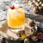 A cocktail of a whiskey sour sits on a small tray surrounded by little quail eggs.
