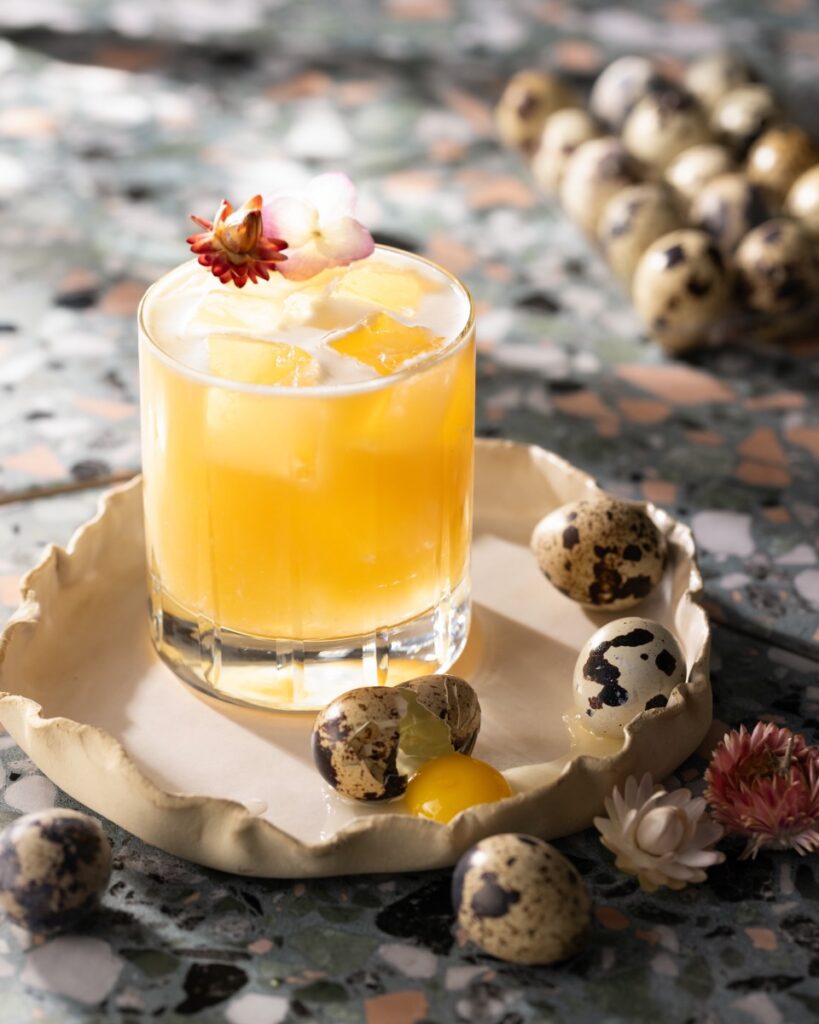 A cocktail of a whiskey sour sits on a small tray surrounded by little quail eggs.