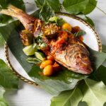 A white plate with leaves every where and a red snapper laying on the plate with taro root smash and salsas.