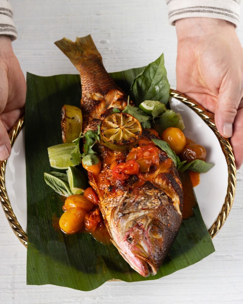 A person presents a white plate with a green banana leaf and a red snapper on top of that.