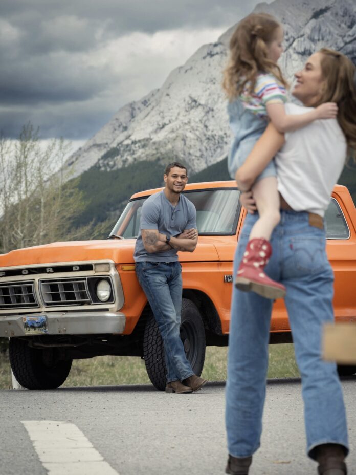 A woman holding a child facing a man leaning on an orange truck with the mountains behind him in the distance.