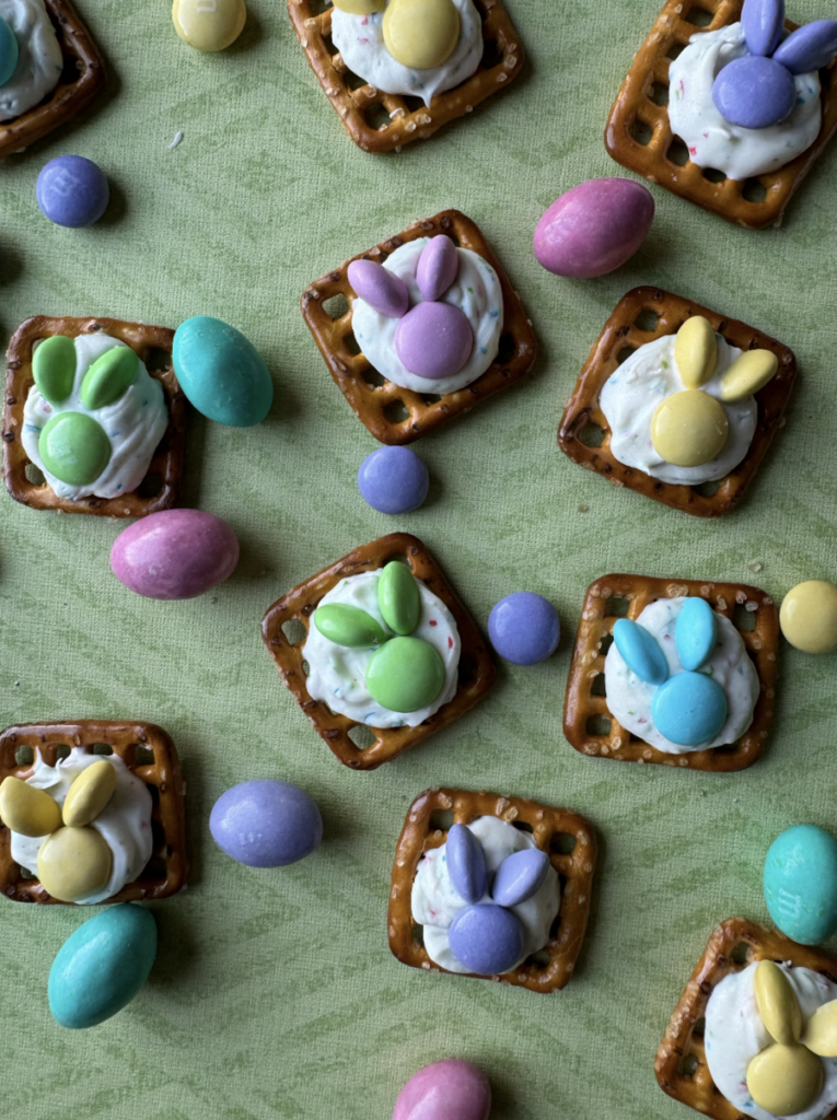 Chocolate Pretzel Easter Bunny Treats sit on a green striped background made out of pretzels, chocolate, and M&Ms with M&Ms tossed between the treats.
