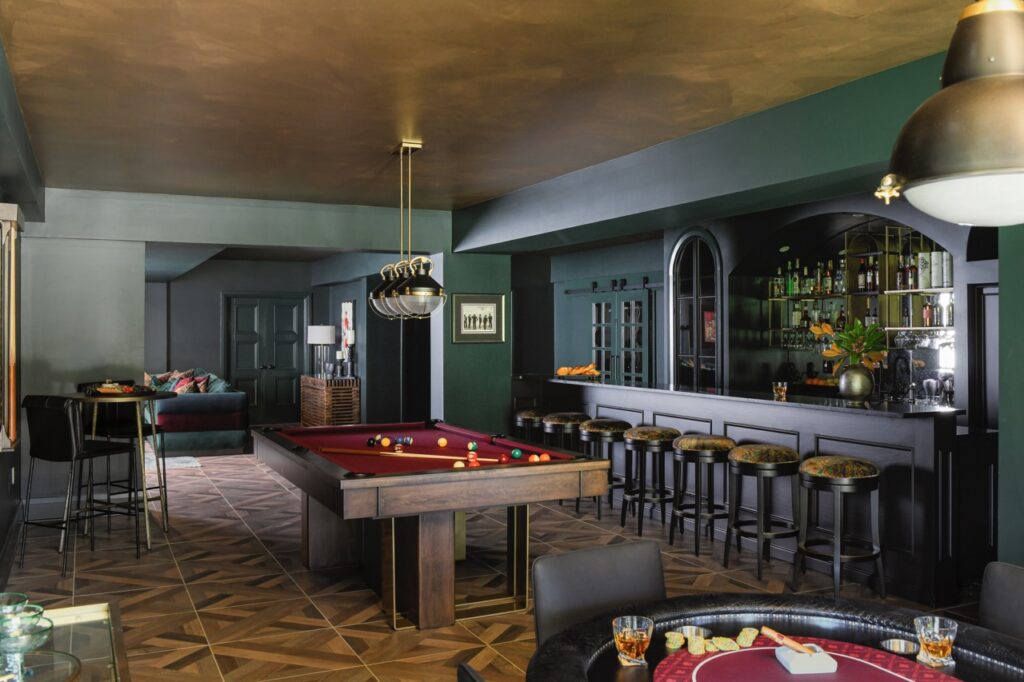 A pool table sits on a pattern carpet in an open bar area featuring stools.