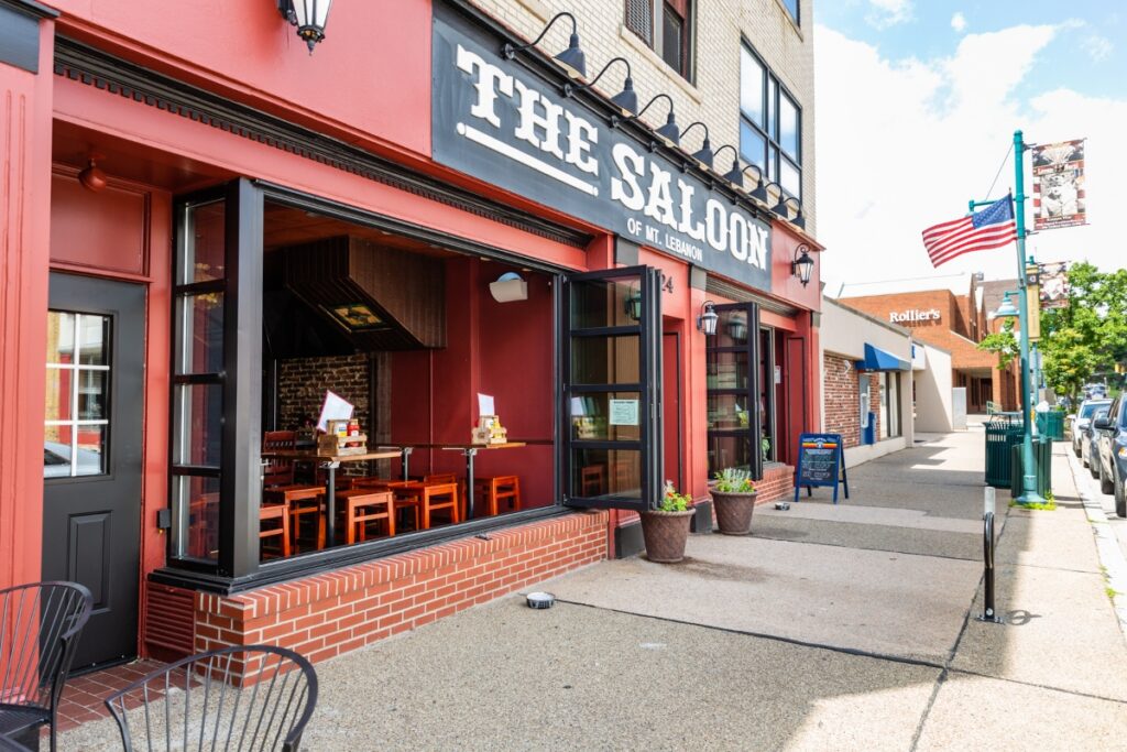 The exterior of The Saloon of Mt. Lebanon on Washington Road, featuring a bright red storefront, open garage-style windows with indoor seating, and an American flag out front.