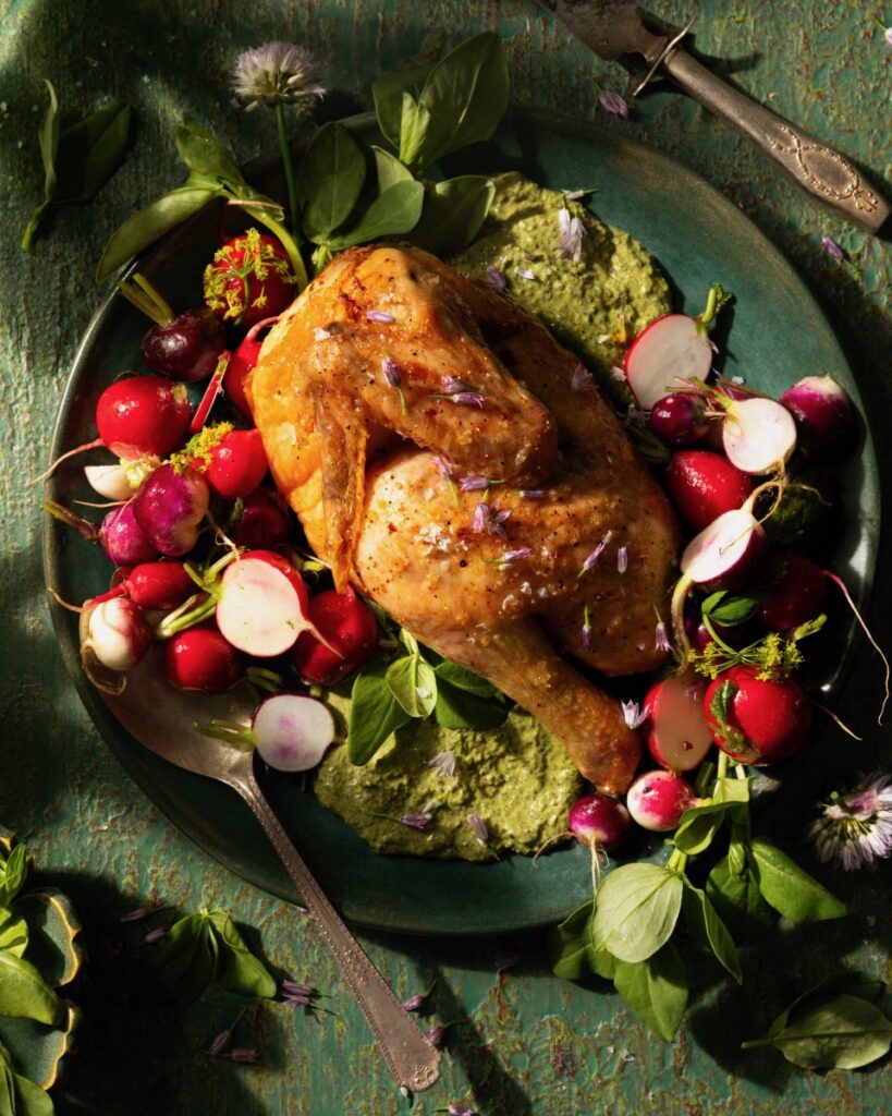 A green plate on a green surface plates spring chicken with a fava shoot pesto and half-cut radishes.