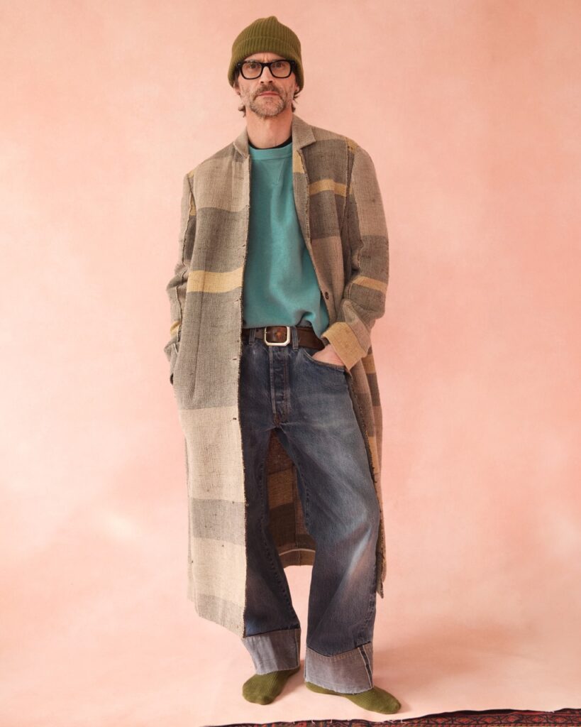 A man in a long woven jacket and beanie stands in front of a pink backdrop.