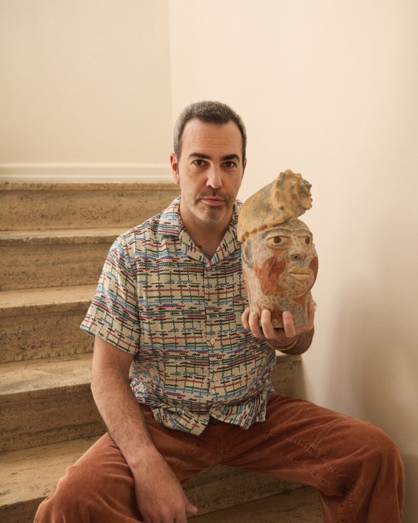 A man in a pattern button up and orange pants sits on a set of stairs holding a vintage face sculpture.