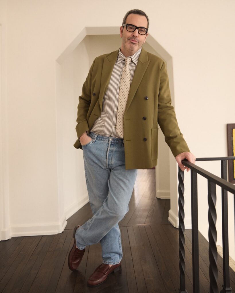A man leans against a railing in a green blazer, white tie, and jeans.
