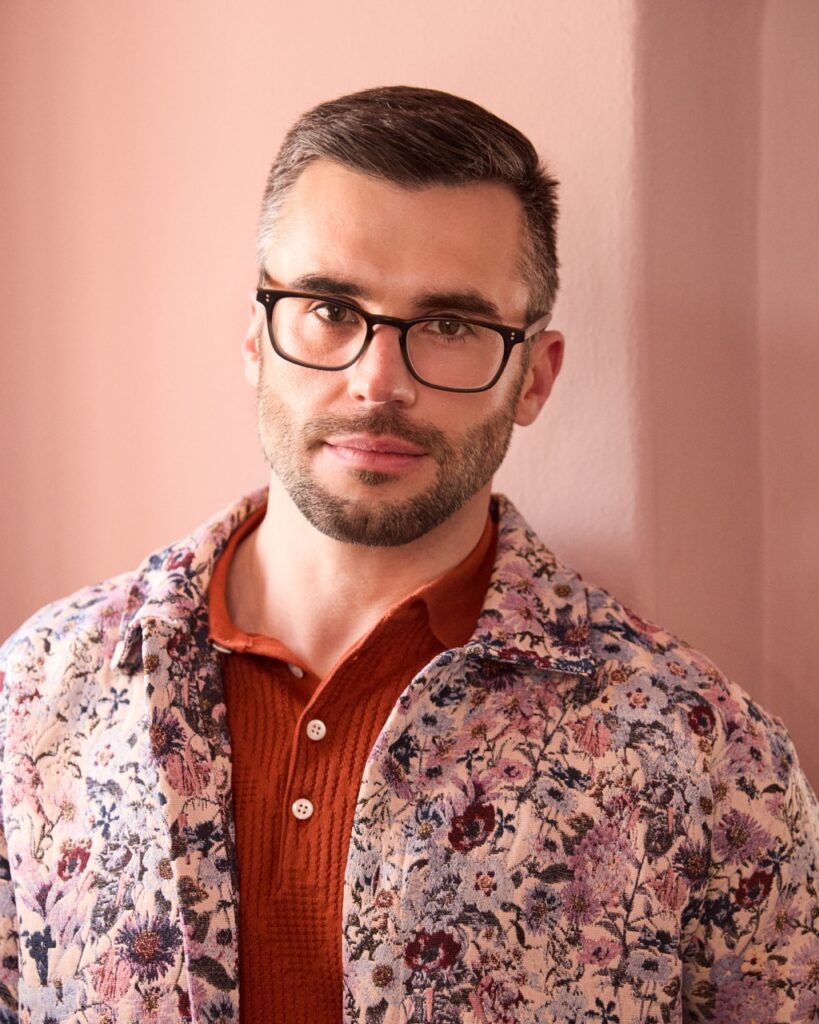 A man stands in an orange polo and floral jacket in front of a pink wall.