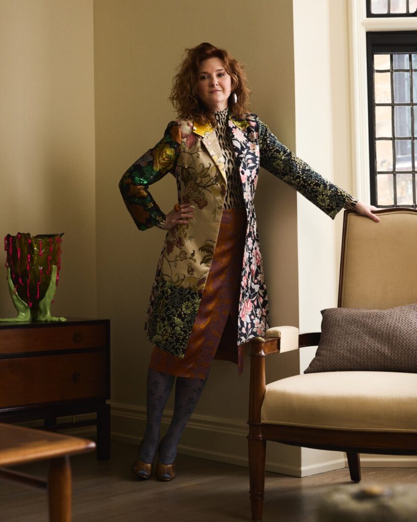 A woman in a long pattern jacket leans her hand on a beige chair.
