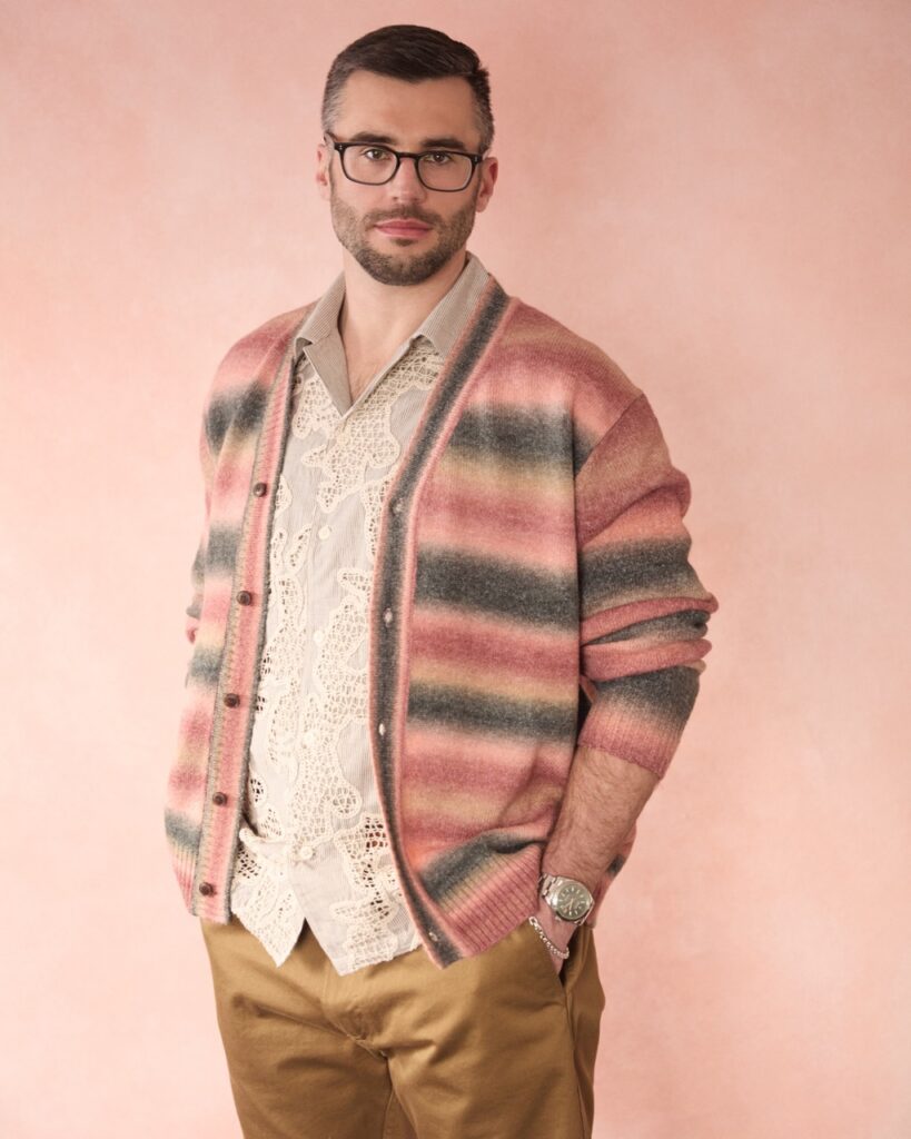 A man stands in a striped sweater and button up with glasses on.