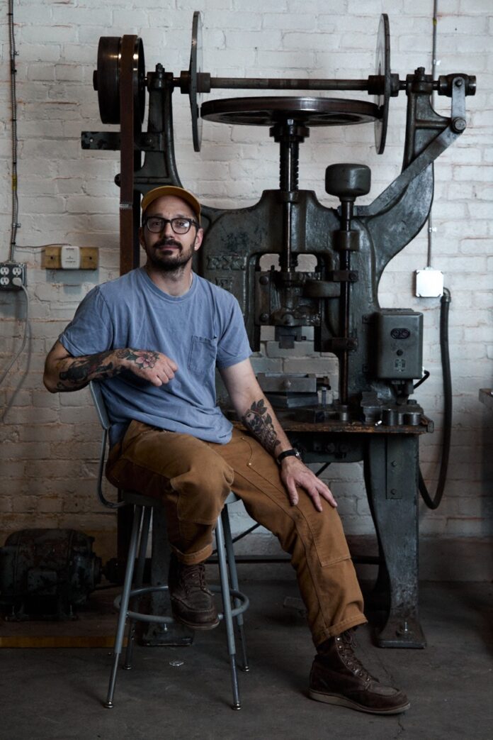 A man in a blue shirt and khakis sits in front of an old metal machine.