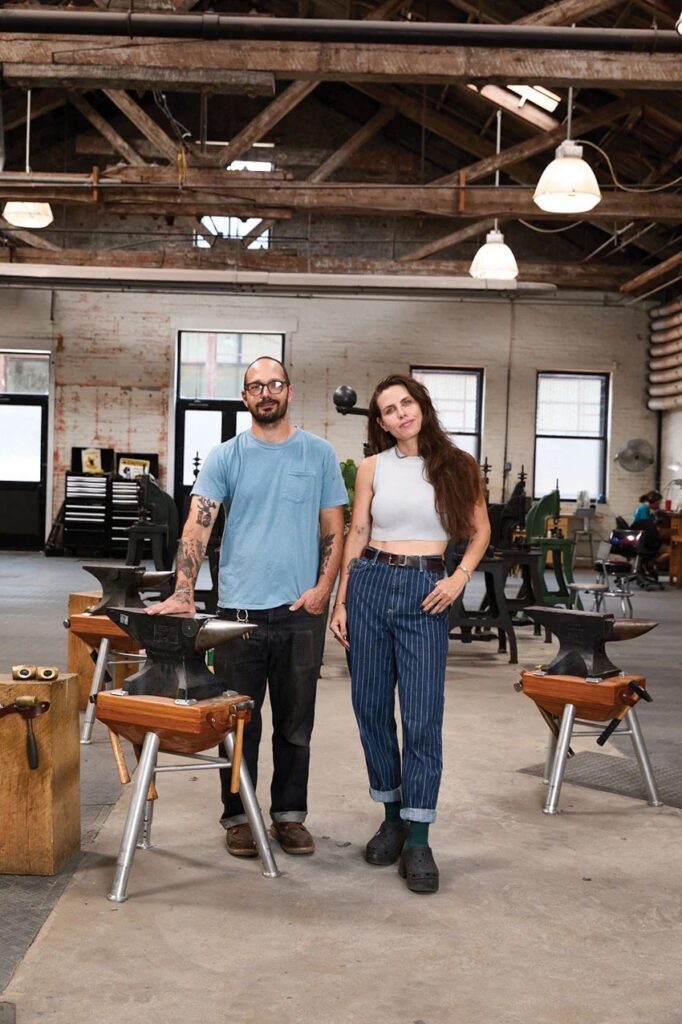 A man in a blue shirt and woman in a white shirt stand in Studebaker Metals Studios with each other around anvils.