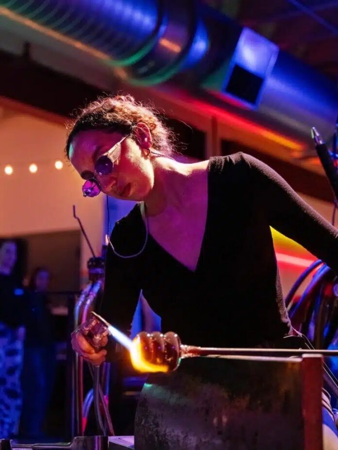 An artist blows glass with goggles on.