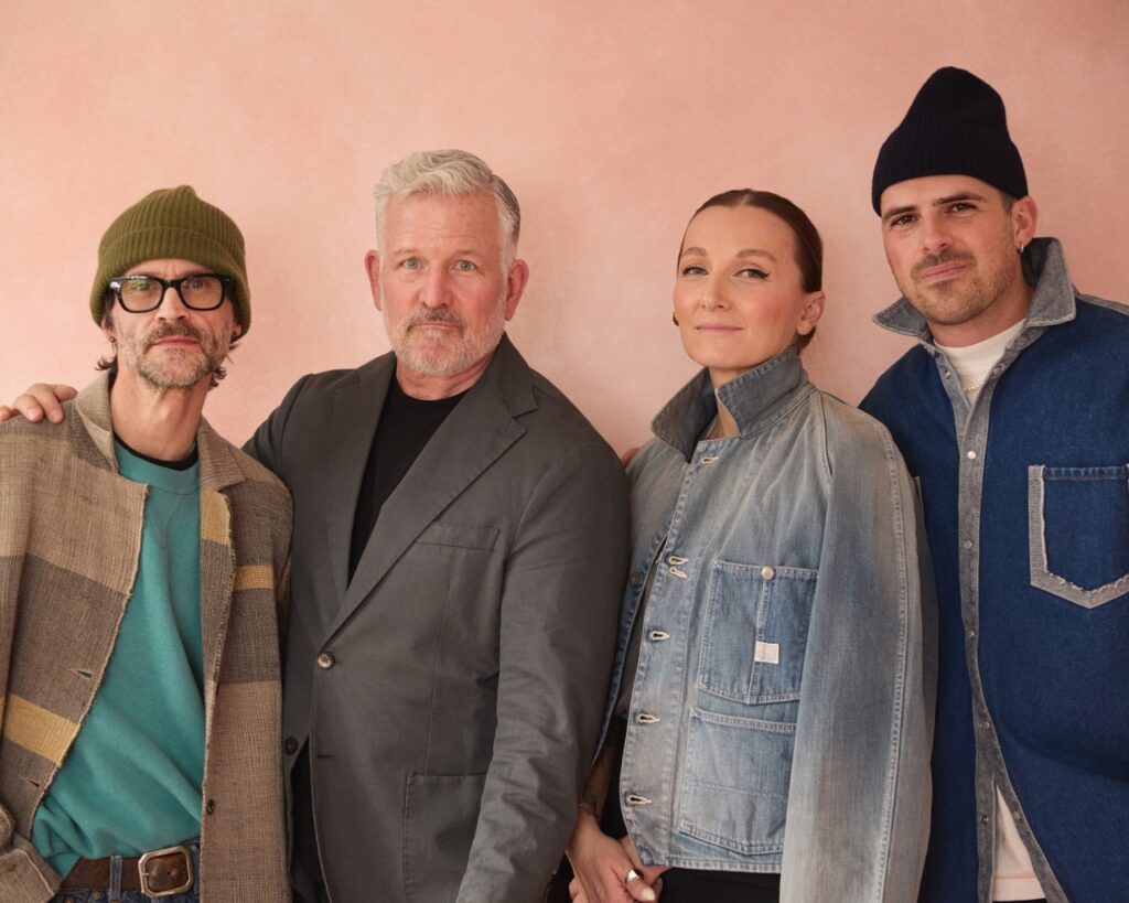 Three men in jackets and a woman in a jean jacket stand together against a pink backdrop.
