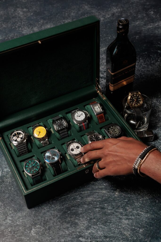 A luxury men’s watch collection from Orr’s Jewelers featuring Breitling, Cartier, Panerai, and IWC timepieces in a green velvet-lined leather display box beside a bottle of Johnnie Walker Double Black.