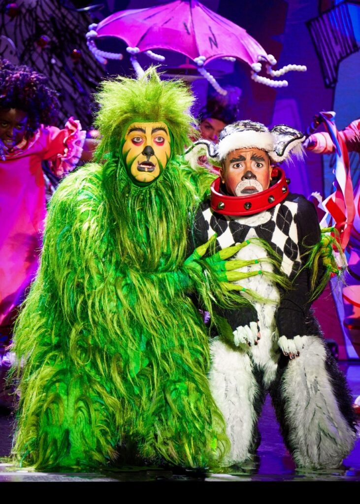 The Grinch in a green fuzzy costume kneels next to Max the dog.