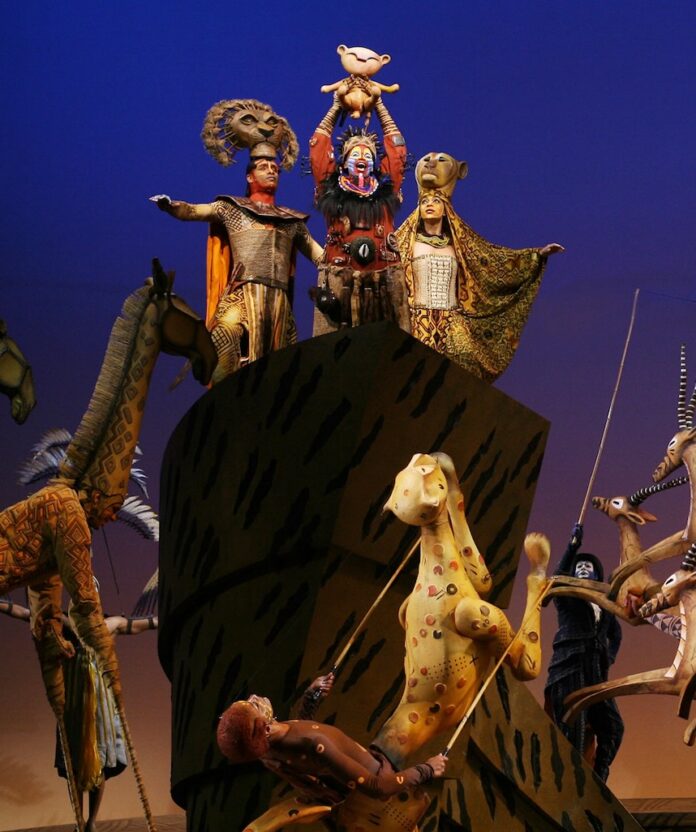 The Lion King on Broadway with three actors on top of a large rock and puppet animals around it.