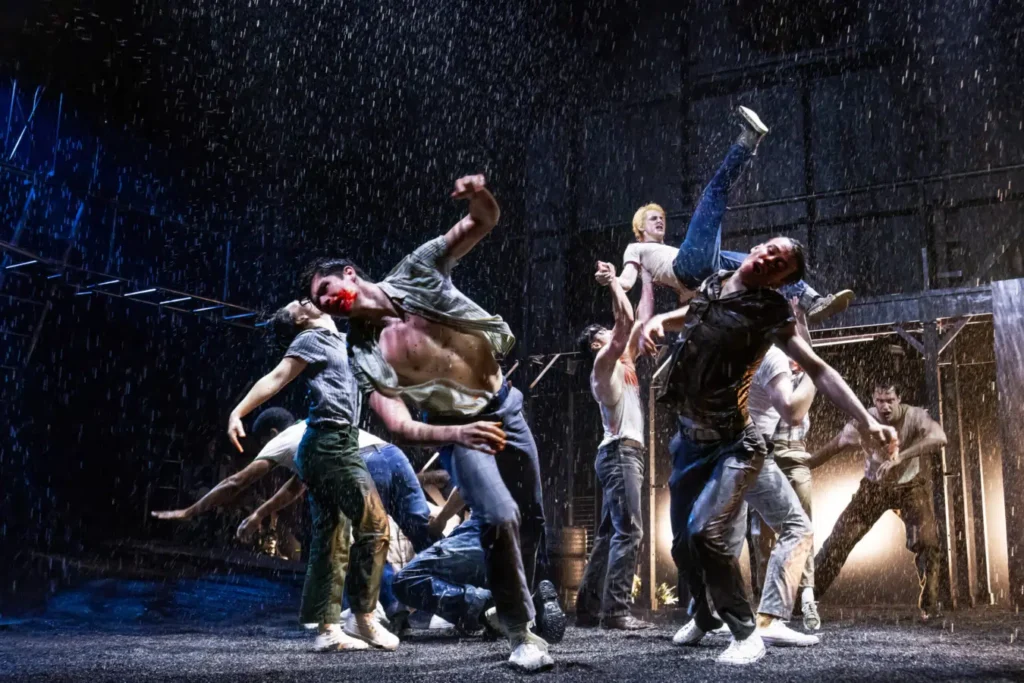 Kids in The Outsiders Broadway musical fake combat fighting on stage in the rain.