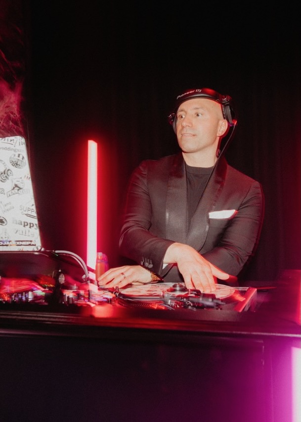 A man DJs at a table with pink and red lights behind him.