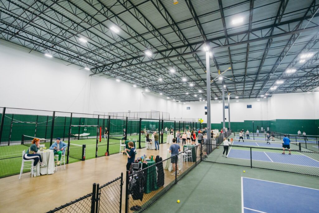 An indoor padel court at Matt’s Pickle and Padel in Sewickley, featuring blue turf, glass walls, and a high-ceiling industrial sports facility.