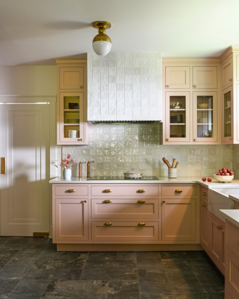 A bright, modern kitchen designed by Michelle Gage featuring blush pink cabinets, a white tiled range hood, gold hardware, and dark stone flooring.