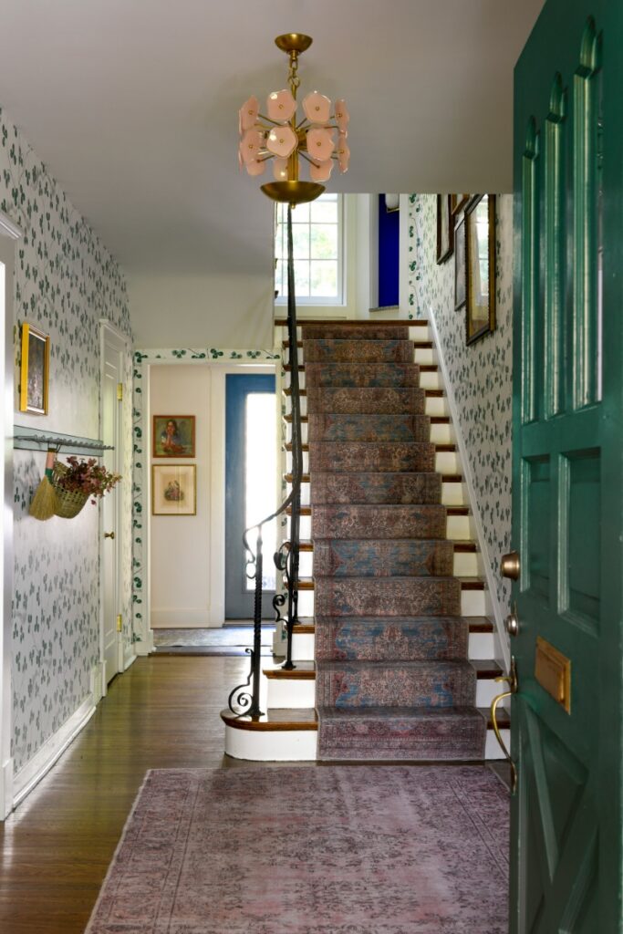 A stylish home entryway featuring Svenskt Tenn clover-patterned wallpaper, a staircase with a vintage-style runner, and a floral-inspired Visual Comfort pendant light.