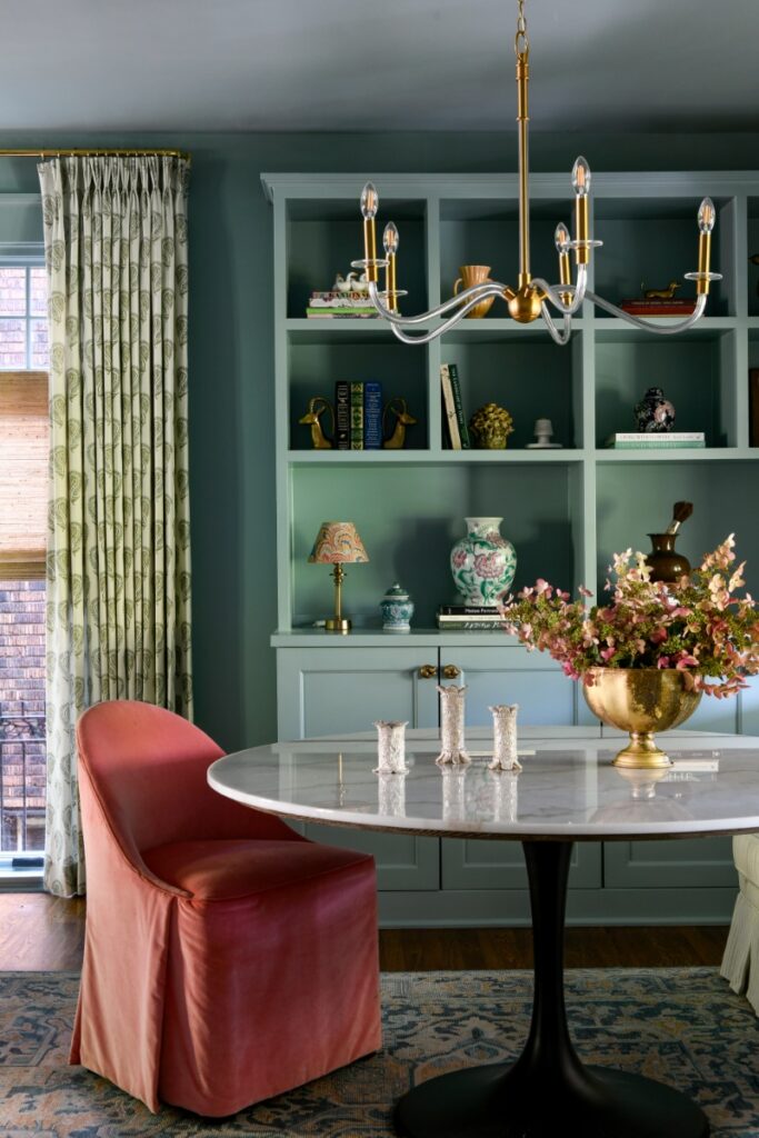 A stylish library or gaming nook featuring teal built-in bookshelves, a round marble-top pedestal table, and a red velvet chair.