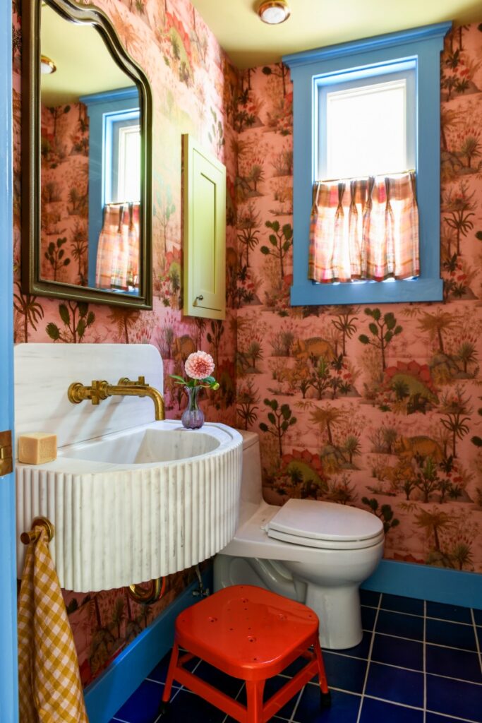 A vibrant powder room designed by Michelle Gage featuring orange themed wallpaper, a fluted marble pedestal sink with brass fixtures, and bold blue window trim.