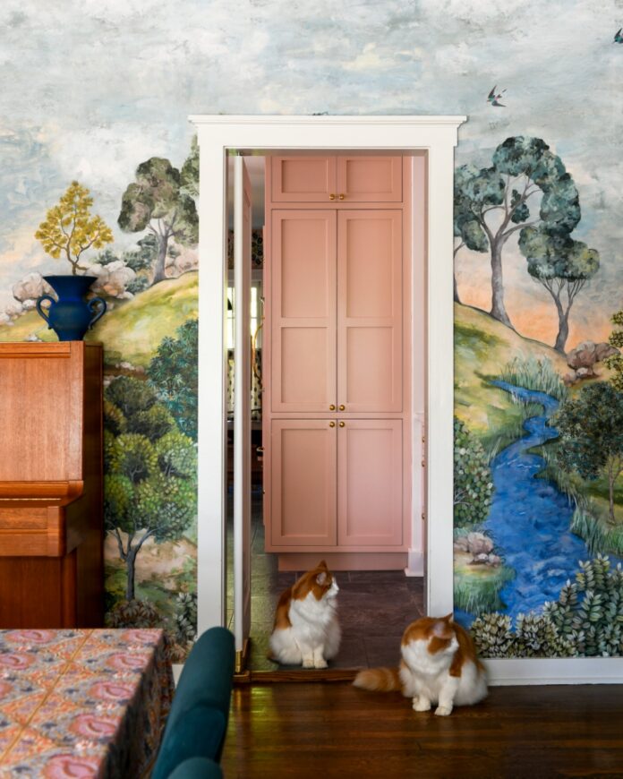 A whimsical dining room featuring a Designers Guild forest landscape mural, two orange and white cats near the doorway to a blush pink kitchen, and a wooden piano.