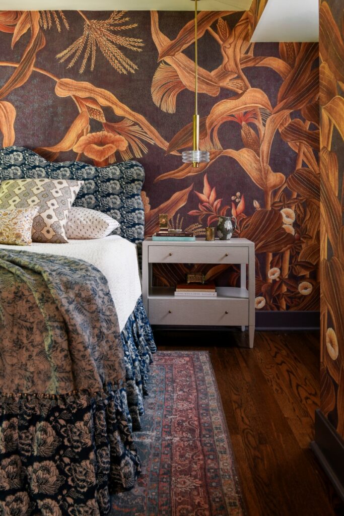 A whimsical bedroom corner featuring a scalloped blue and white floral headboard, a gray textured nightstand, and bold aubergine Iksel cornflower wallpaper.